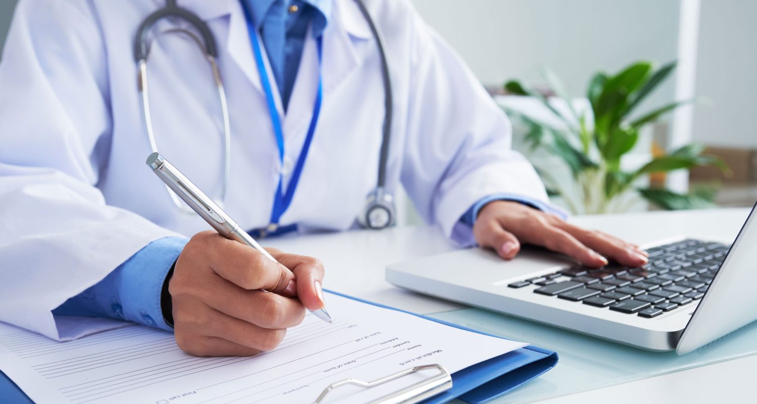 hands-unrecognizable-female-doctor-writing-form-typing-laptop-keyboard